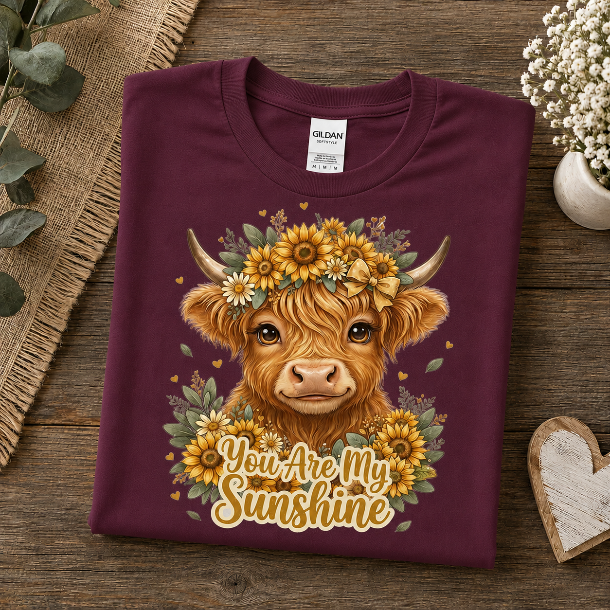 A folded maroon Gildan t-shirt with a detailed graphic of a Highland cow wearing a sunflower crown and the text 'You Are My Sunshine', displayed on a rustic wooden background with decorative props.