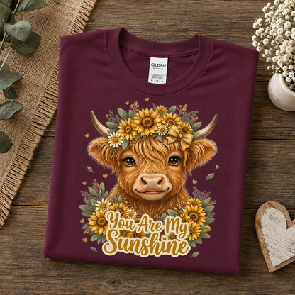 A folded maroon Gildan t-shirt with a detailed graphic of a Highland cow wearing a sunflower crown and the text 'You Are My Sunshine', displayed on a rustic wooden background with decorative props.