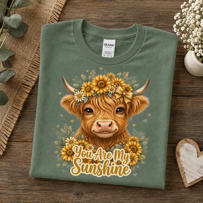 A folded military green Gildan t-shirt with a graphic of a Highland cow wearing a sunflower crown and the text 'You Are My Sunshine', displayed on a rustic wooden background with decorative props.