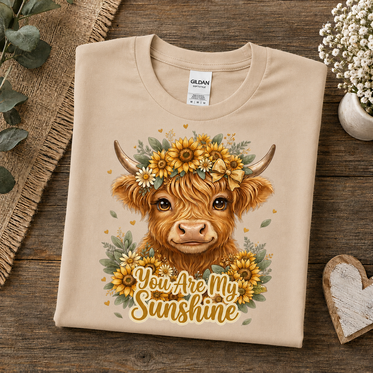 A sand colored Gildan t-shirt is neatly folded on a rustic wooden surface, decorated with eucalyptus leaves, a small vase of white flowers, and a wooden heart. The shirt features a graphic of a Highland cow wearing a yellow sunflower crown with the text 'You Are My Sunshine' written below in a warm yellow font.