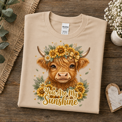 A sand colored Gildan t-shirt is neatly folded on a rustic wooden surface, decorated with eucalyptus leaves, a small vase of white flowers, and a wooden heart. The shirt features a graphic of a Highland cow wearing a yellow sunflower crown with the text 'You Are My Sunshine' written below in a warm yellow font.