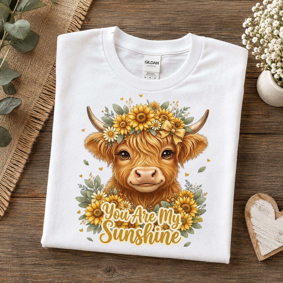 A folded white Gildan t-shirt with a graphic of a Highland cow wearing a sunflower crown and the text 'You Are My Sunshine', displayed on a rustic wooden background with decorative props.