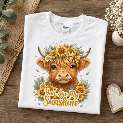 A folded white Gildan t-shirt with a graphic of a Highland cow wearing a sunflower crown and the text 'You Are My Sunshine', displayed on a rustic wooden background with decorative props.