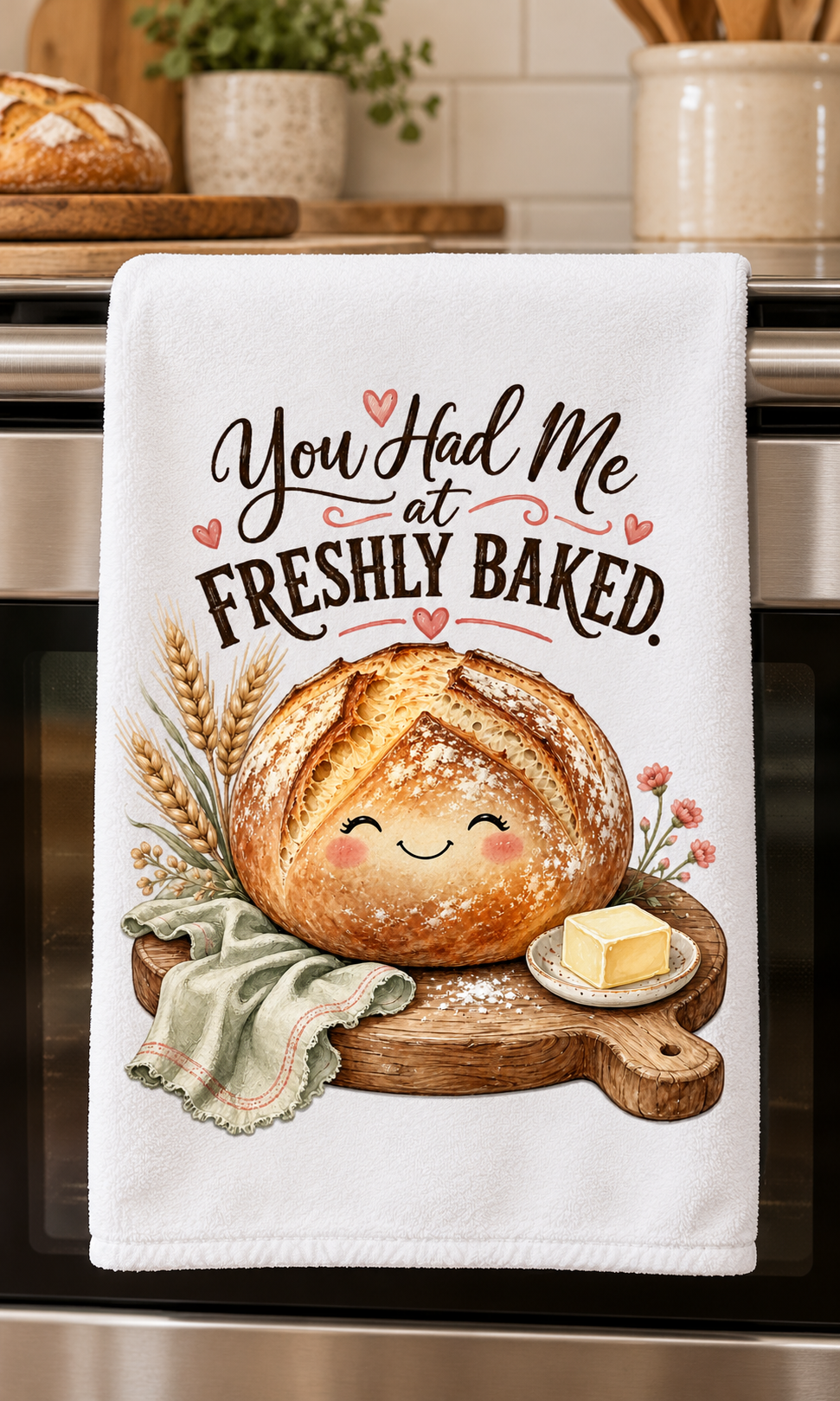 A white kitchen towel hanging on a stainless steel oven handle, featuring a cute graphic design of a smiling round loaf of bread on a wooden board with wheat stalks, a green cloth, and a pat of butter. The text reads, 'You Had Me at FRESHLY BAKED.