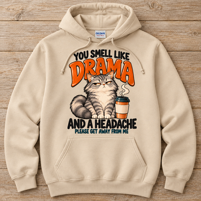 A sand pullover hoodie laid flat on a rustic wood background, featuring a graphic print of a grumpy cat next to a coffee cup with the text 'YOU SMELL LIKE DRAMA AND A HEADACHE PLEASE GET AWAY FROM ME'. 