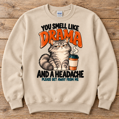 A sand crewneck sweatshirt with a humorous graphic print featuring a grumpy cat, a coffee cup, and the text 'YOU SMELL LIKE DRAMA AND A HEADACHE PLEASE GET AWAY FROM ME' in black and orange lettering.