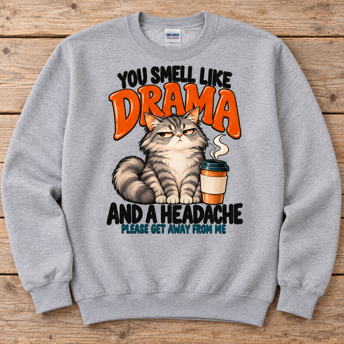 A light gray crewneck sweatshirt featuring a humorous graphic print of a grumpy cat next to a coffee cup, with the text 'YOU SMELL LIKE DRAMA AND A HEADACHE PLEASE GET AWAY FROM ME'. 