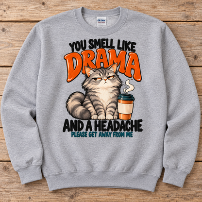A light gray crewneck sweatshirt featuring a humorous graphic print of a grumpy cat next to a coffee cup, with the text 'YOU SMELL LIKE DRAMA AND A HEADACHE PLEASE GET AWAY FROM ME'. 