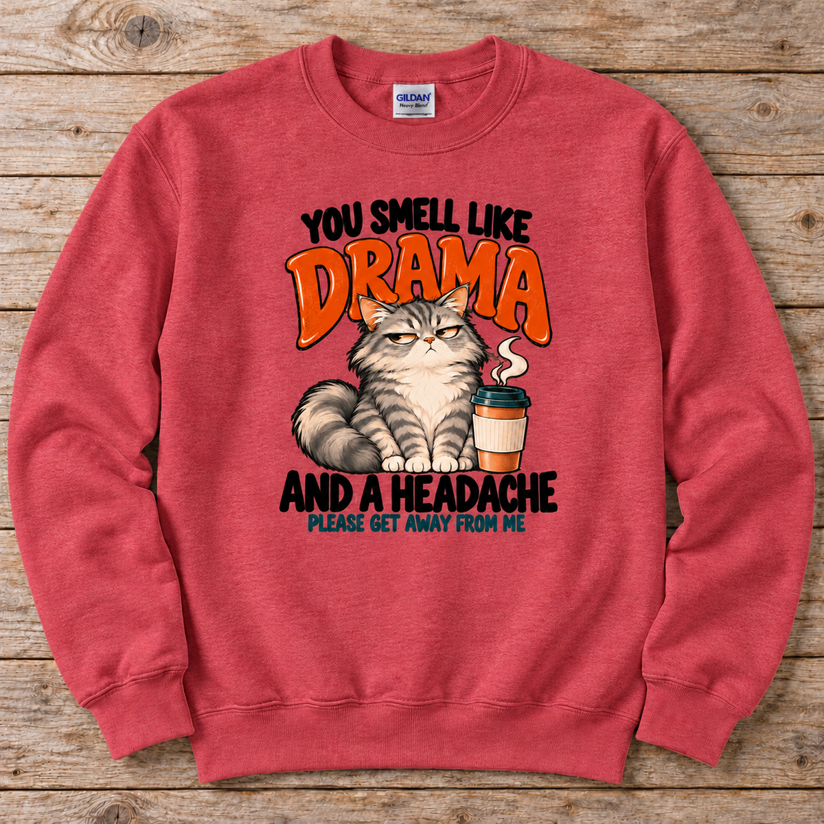 A heather carlet red crewneck sweatshirt featuring a humorous graphic print of a grumpy cat next to a coffee cup, with the text 'YOU SMELL LIKE DRAMA AND A HEADACHE PLEASE GET AWAY FROM ME'. 