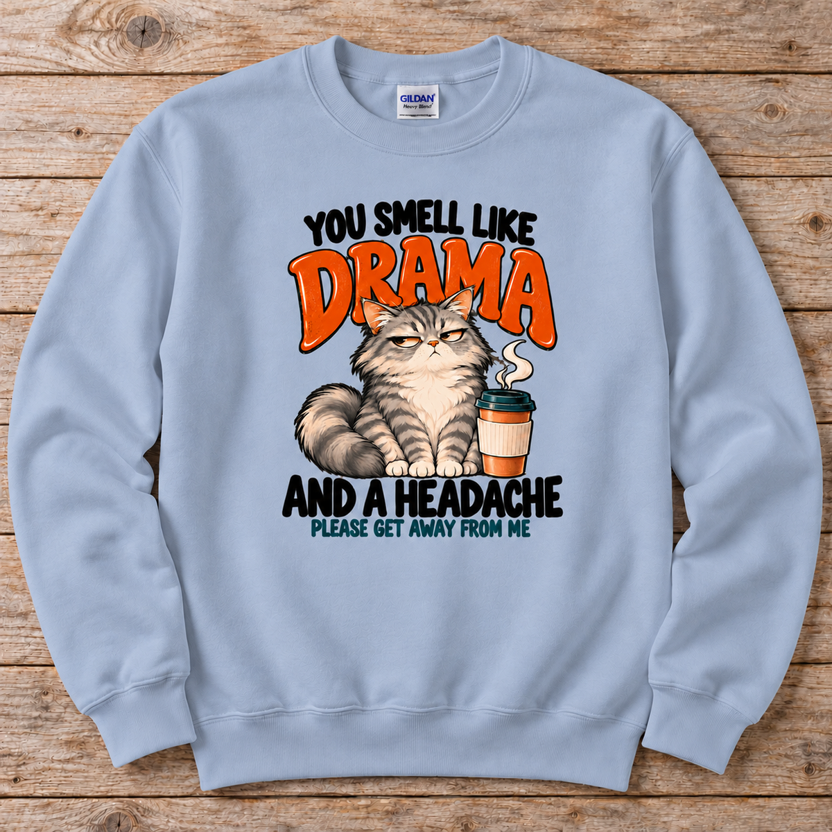 A light blue crewneck sweatshirt featuring a humorous graphic print of a grumpy cat next to a coffee cup, with the text 'YOU SMELL LIKE DRAMA AND A HEADACHE PLEASE GET AWAY FROM ME'. 