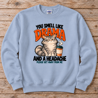A light blue crewneck sweatshirt featuring a humorous graphic print of a grumpy cat next to a coffee cup, with the text 'YOU SMELL LIKE DRAMA AND A HEADACHE PLEASE GET AWAY FROM ME'. 
