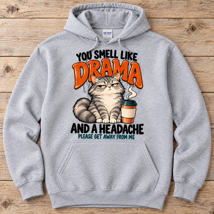 A light gray heather hoodie laid flat on a rustic wooden background. The hoodie features a graphic print of a grumpy tabby cat next to a coffee cup, with the text 'YOU SMELL LIKE DRAMA AND A HEADACHE PLEASE GET AWAY FROM ME' in black and orange lettering.