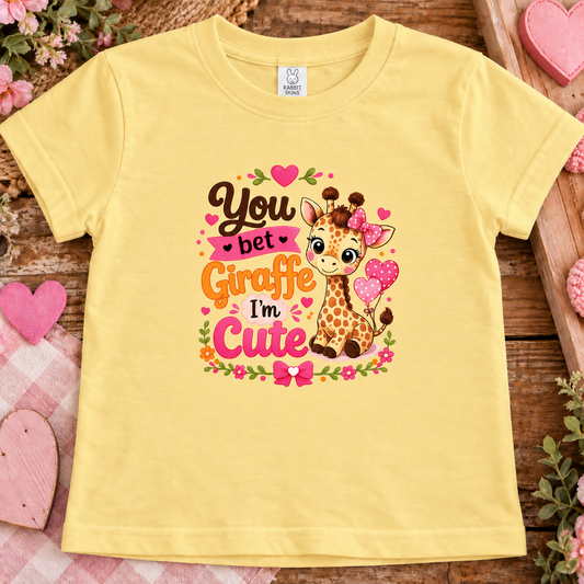 A butter yellow short-sleeve kids' t-shirt with a colorful graphic of a cartoon giraffe and the text 'You bet Giraffe I'm Cute' in pink, orange, and brown lettering. The t-shirt is displayed flat on a rustic wooden surface decorated with pink hearts, flowers, and a pink plaid blanket.