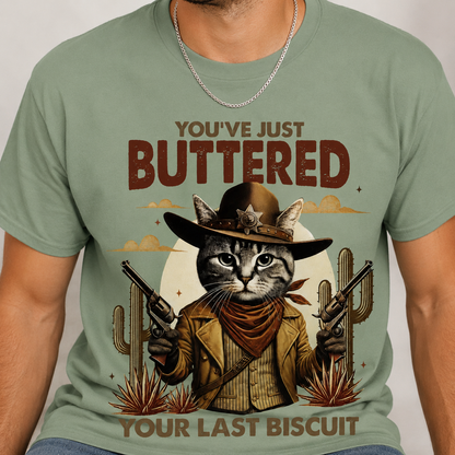 Bay colored t-shirt with a graphic of a cat in a cowboy outfit and text 'You've Just Buttered Your Last Biscuit' on a plain background.