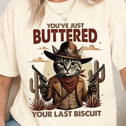 Ivory colored T-shirt with a graphic of a cat in a cowboy outfit and text 'You've just buttered your last biscuit' on a beige background.