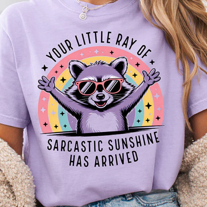 Sarcastic raccoon with arms wide open wearing sunglasses in front of a rainbow design on a Orchid Comfort Colors Shirt