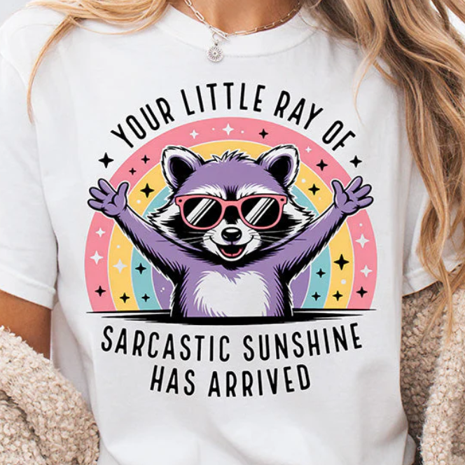 Sarcastic raccoon with arms wide open wearing sunglasses in front of a rainbow design on a White Comfort Colors Shirt