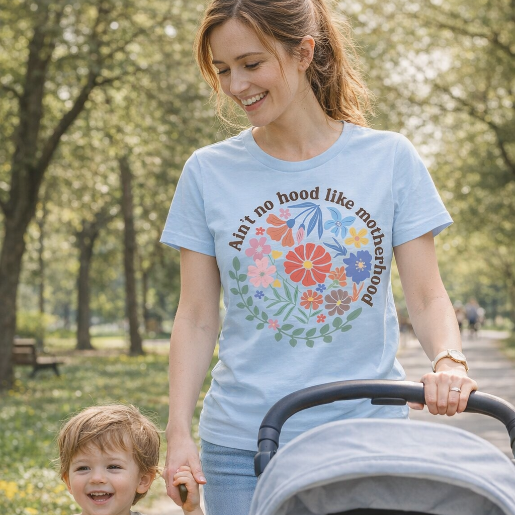 Woman walking with a child and stroller in a park, wearing a light blue t-shirt with floral design and text.