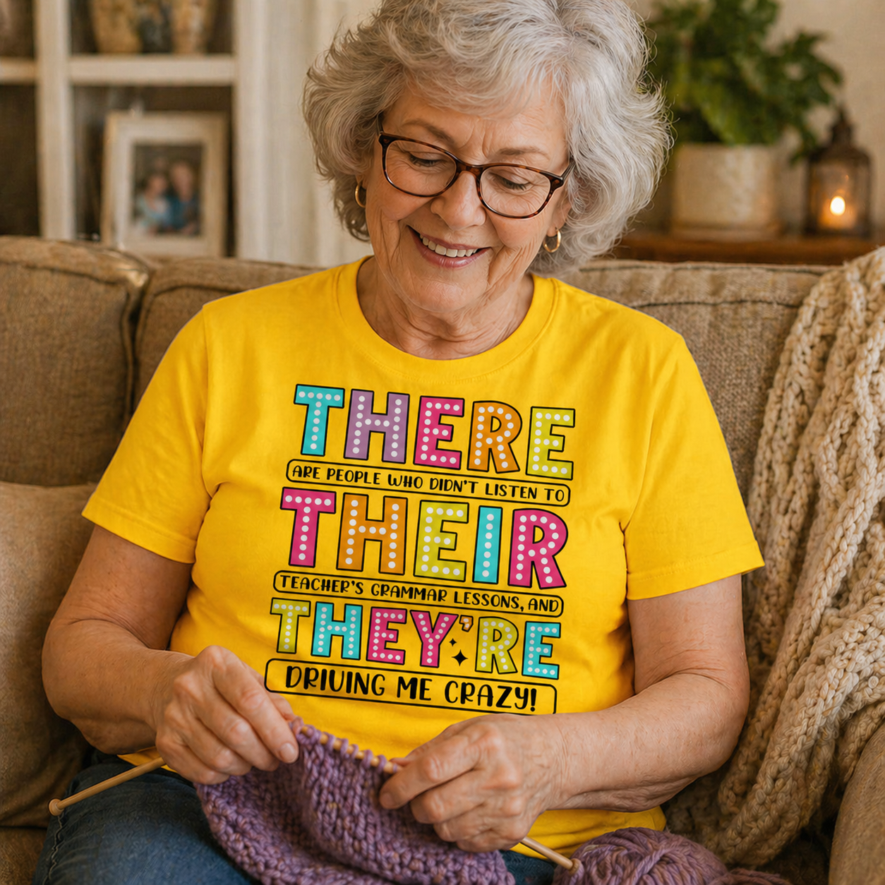 Woman sitting on a couch wearing a daisy colored  t-shirt with colorful text, knitting a purple item.