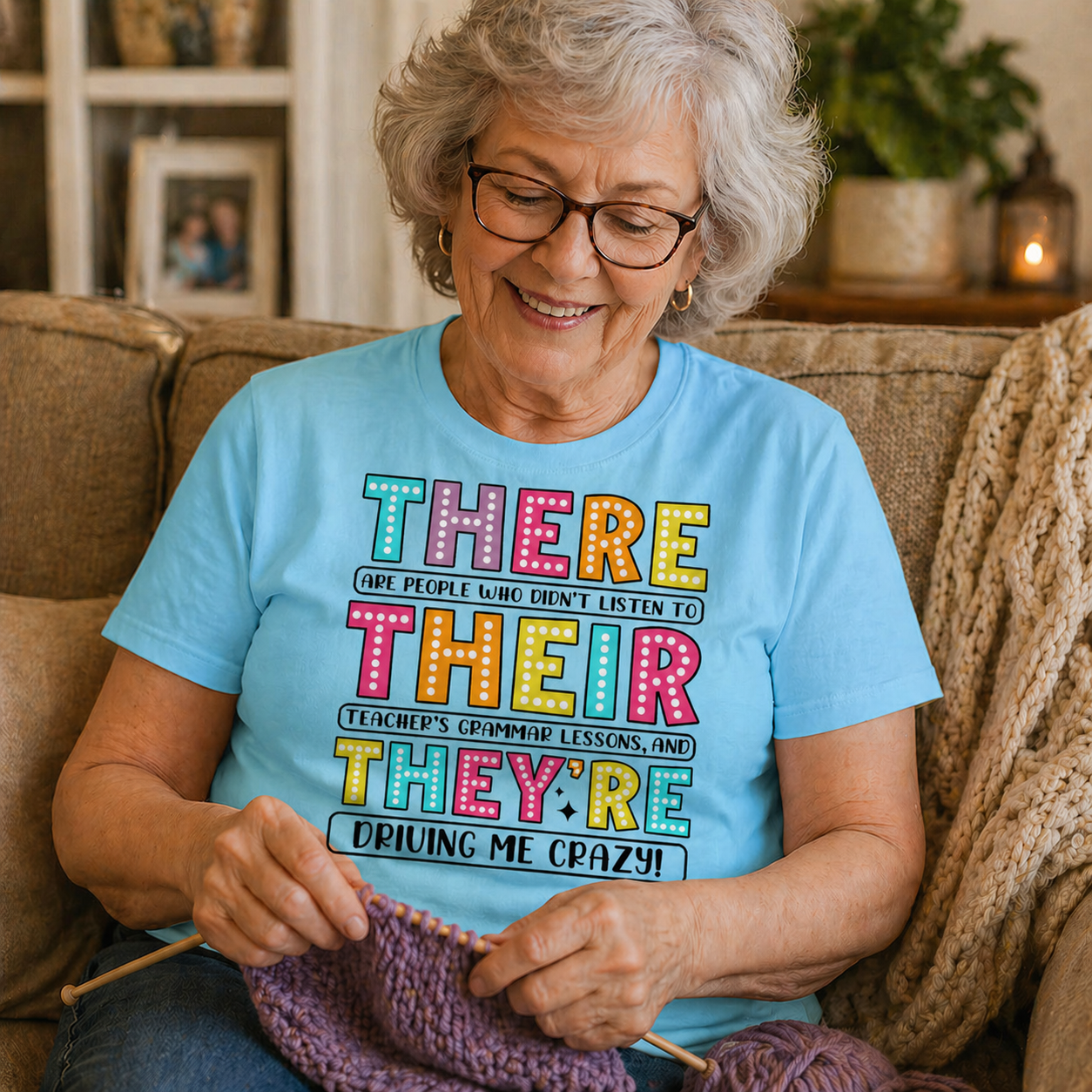 Woman sitting on a couch wearing a sky blue t-shirt with colorful text, knitting a purple item.