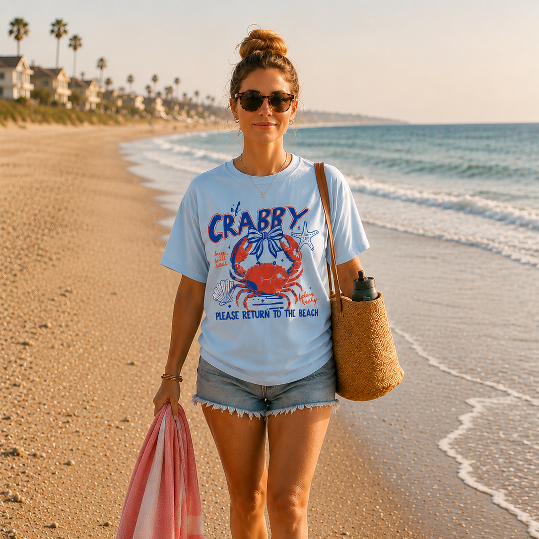 Woman walking barefoot on the beach wearing a light blue graphic t-shirt featuring a retro red and blue crab design with the phrase “If CRABBY Please Return To The Beach.”