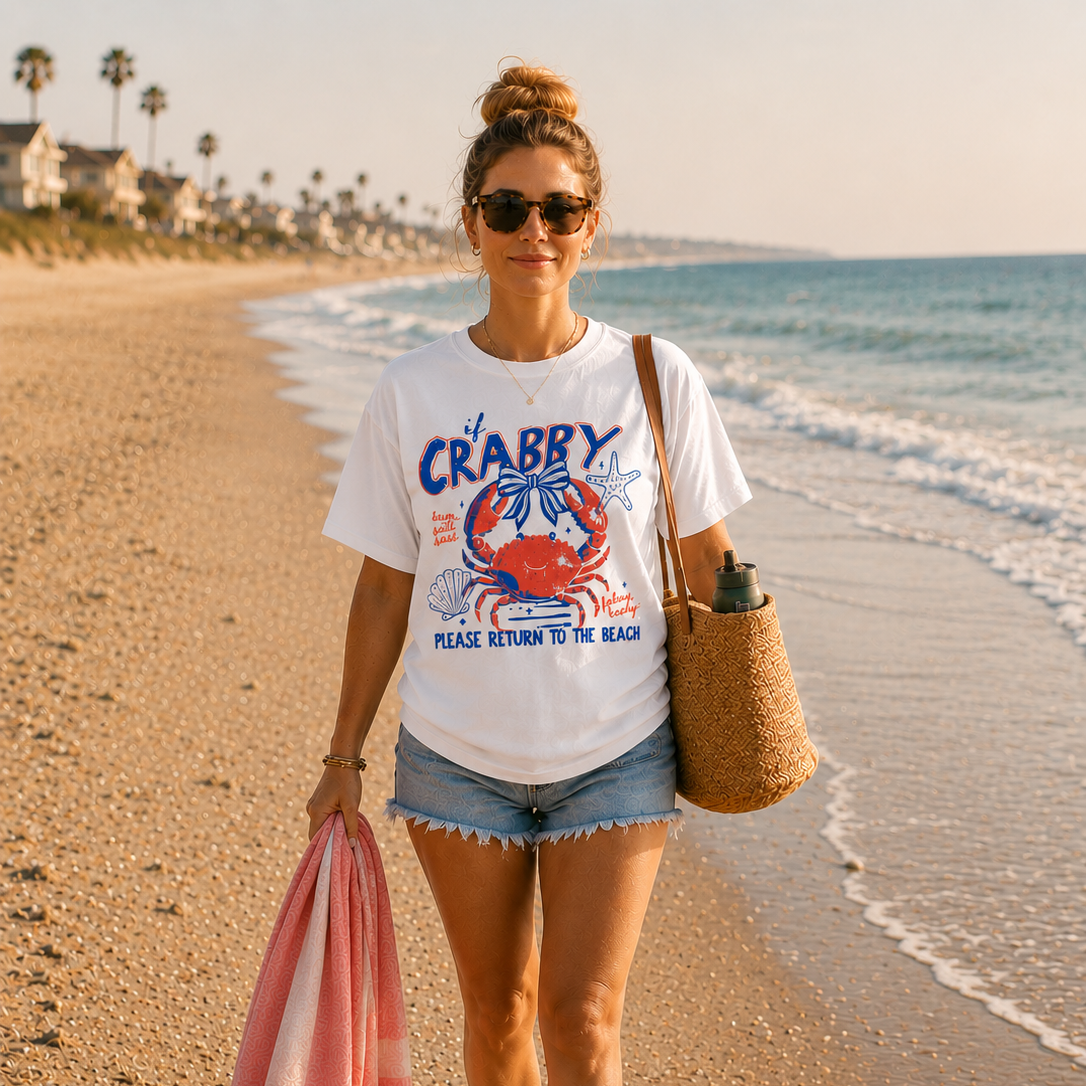 Woman walking barefoot on the beach wearing a white graphic t-shirt featuring a retro red and blue crab design with the phrase “If CRABBY Please Return To The Beach.”