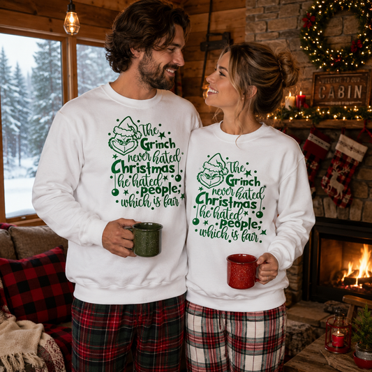 A man and a woman standing in a cozy living room wearing matching white crewneck sweatshirts with a green graphic that reads 'The Grinch never hated Christmas he hated people, which is fair.' The man wears green plaid pajama pants and holds a dark green mug, while the woman wears red and white plaid pajama pants and holds a red mug.