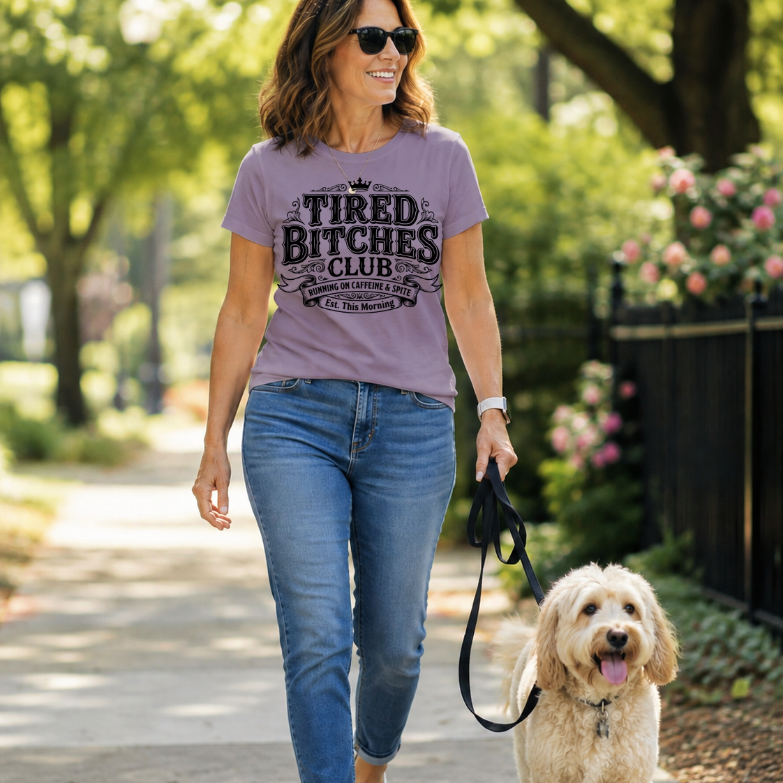A woman walking a fluffy cream-colored dog on a paved path in a park, wearing a lavender graphic t-shirt that reads 'TIRED BITCHES CLUB' in black text, paired with blue jeans and white sneakers.