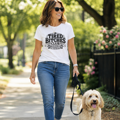 A woman walking a fluffy cream-colored dog on a paved path outdoors, wearing a white graphic t-shirt that reads 'TIRED BITCHES CLUB' in black lettering, paired with blue jeans and white sneakers.