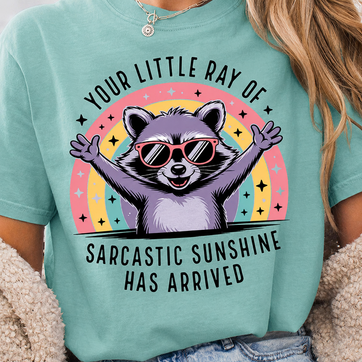 Sarcastic raccoon with arms wide open wearing sunglasses in front of a rainbow design on a Chalky Mint Comfort Colors Shirt
